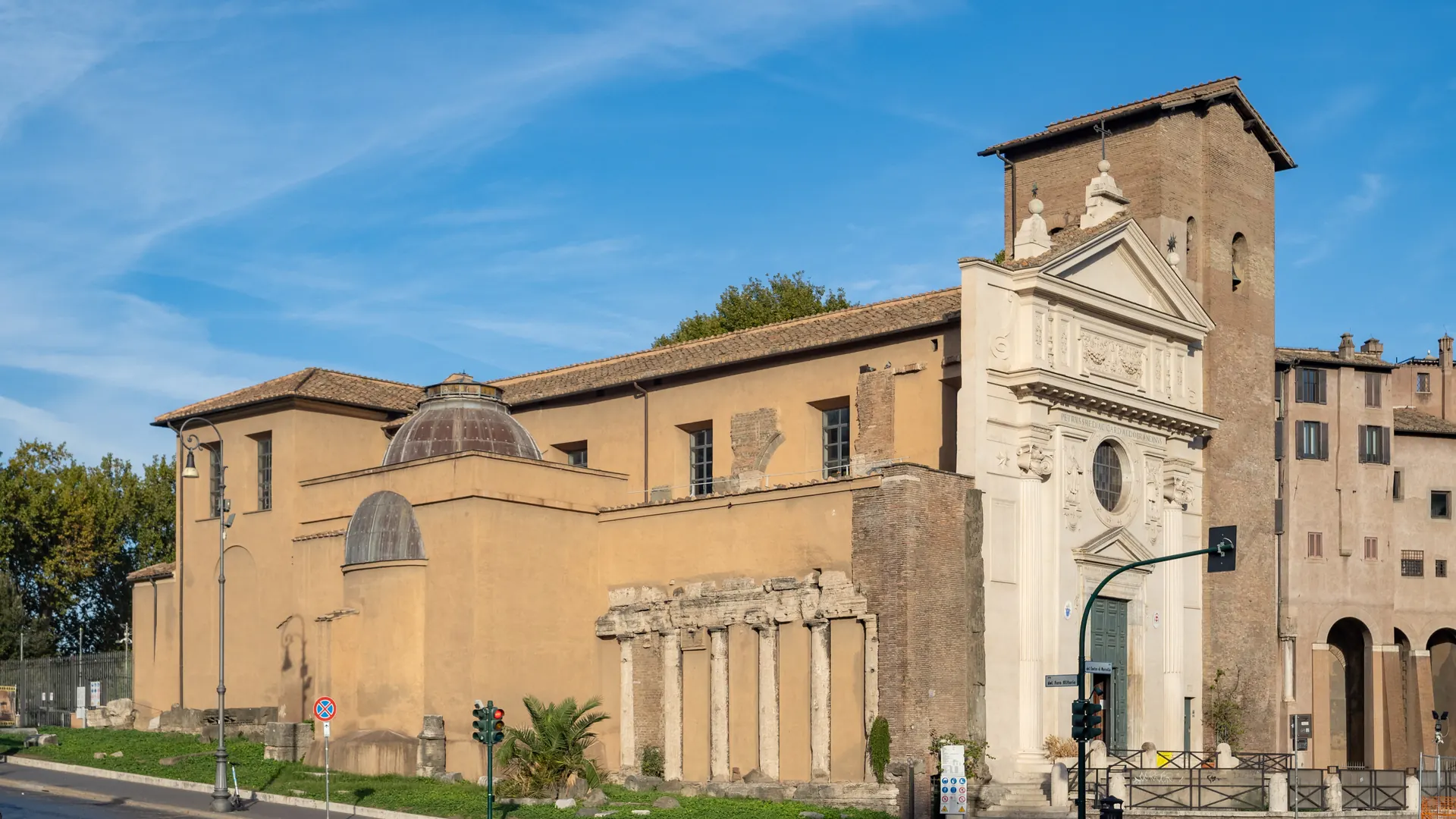 The Basilica of San Nicola in Carcere in Rome in evening light: a pale baroque façade, a medieval brick bell tower and, beside the church, a row of ancient temple columns built into its wall.