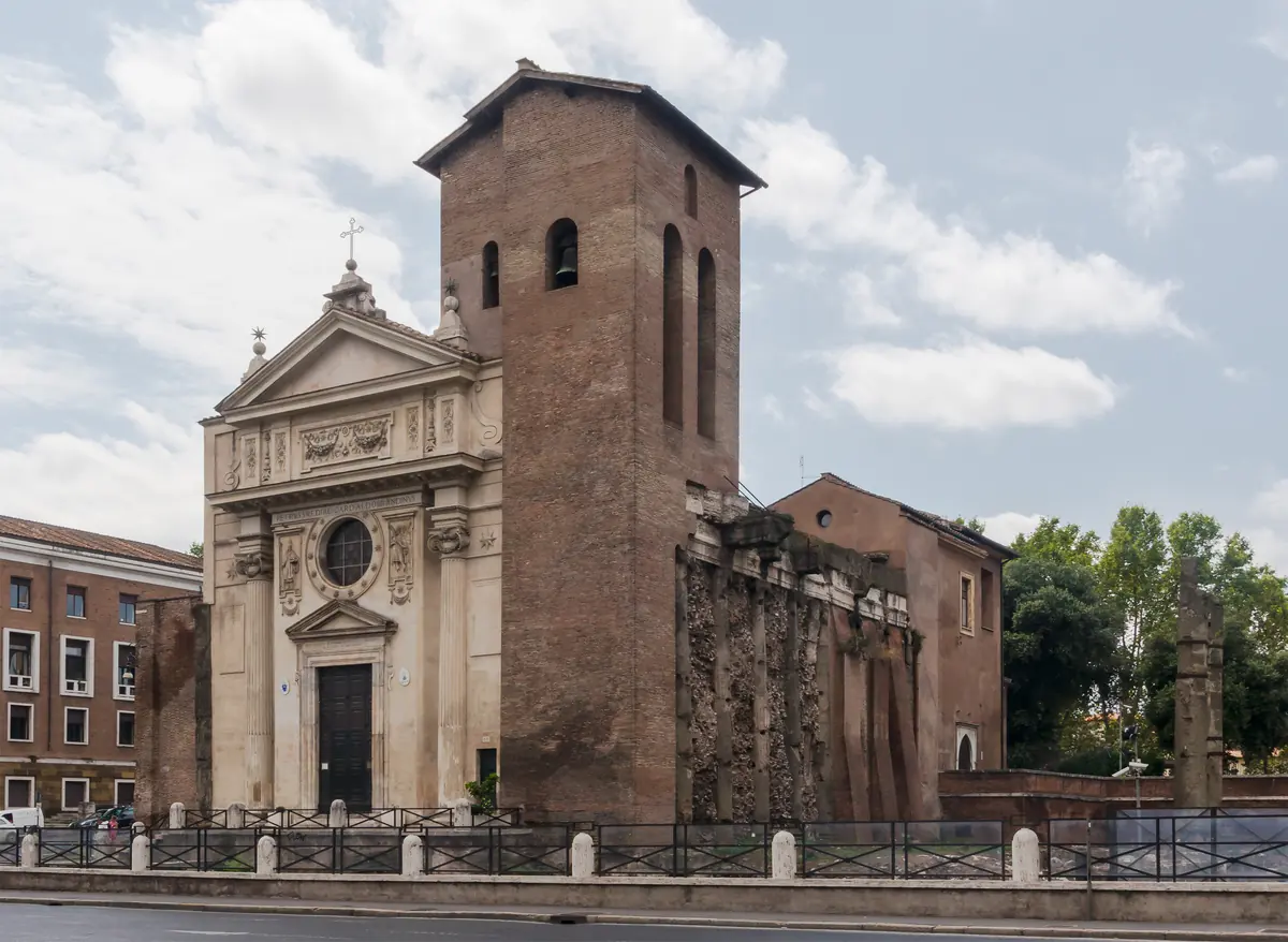 The pale baroque façade of San Nicola in Carcere beside its tall brick medieval bell tower under a cloudy sky
