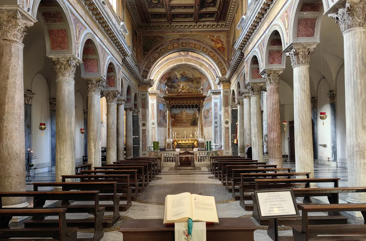 The nave of San Nicola in Carcere seen from the entrance: a long colonnade of ancient columns on both sides, round arches, a coffered ceiling and wooden pews leading to a canopied altar